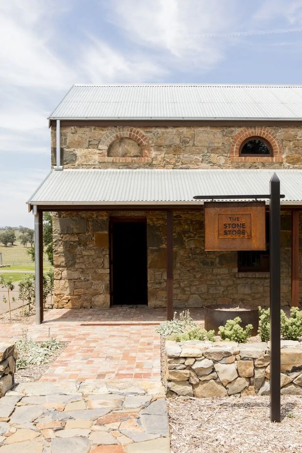 Exterior view of a rustic stone building identified by a timber sign as “The Stone Store.” The building has rough natural-stone walls, arched brick detailing around windows and a sealed opening, corrugated metal roofing with broad verandahs, timber posts, and brick and irregular stone paving. A rural landscape is visible beyond the building.