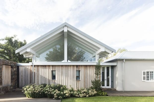 Exterior view of a bright single-storey residential building with a prominent white timber gable roof and exposed rafters. A large triangular glazed gable sits above a vertical timber-clad wall, connecting to a simpler pale rendered wing with white-framed doors and windows. A narrow timber deck, planted garden border, and boundary fencing complete the scene.