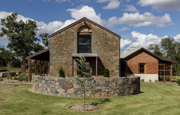 Exterior view of a rural, barn-like residence composed of a central gabled stone volume with a tall arched window, flanked by lower brick gabled wings and open-sided covered verandas. A low stone retaining wall and landscaped lawn sit in the foreground under a bright partly cloudy sky.