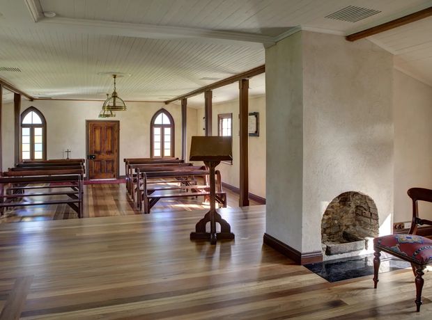 Interior view of a historic chapel or small church with rows of dark timber pews, arched windows, a timber entrance door, exposed dark wood trim and posts, and a small rustic stone fireplace in the foreground. The room has a white painted wood-panel ceiling, warm timber flooring, and traditional ecclesiastical detailing.