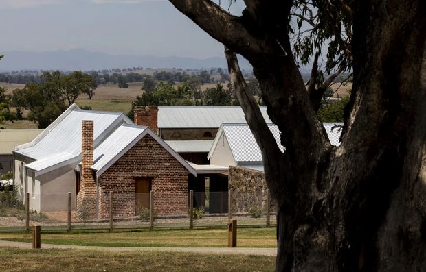 A rural homestead or group of converted farm buildings viewed from a landscaped foreground. The main building has red brick gabled volumes, pale rendered walls, light grey corrugated metal pitched roofs, brick chimneys, and a stone wall section. A large mature tree dominates the right foreground, with open agricultural fields and distant hills beyond.