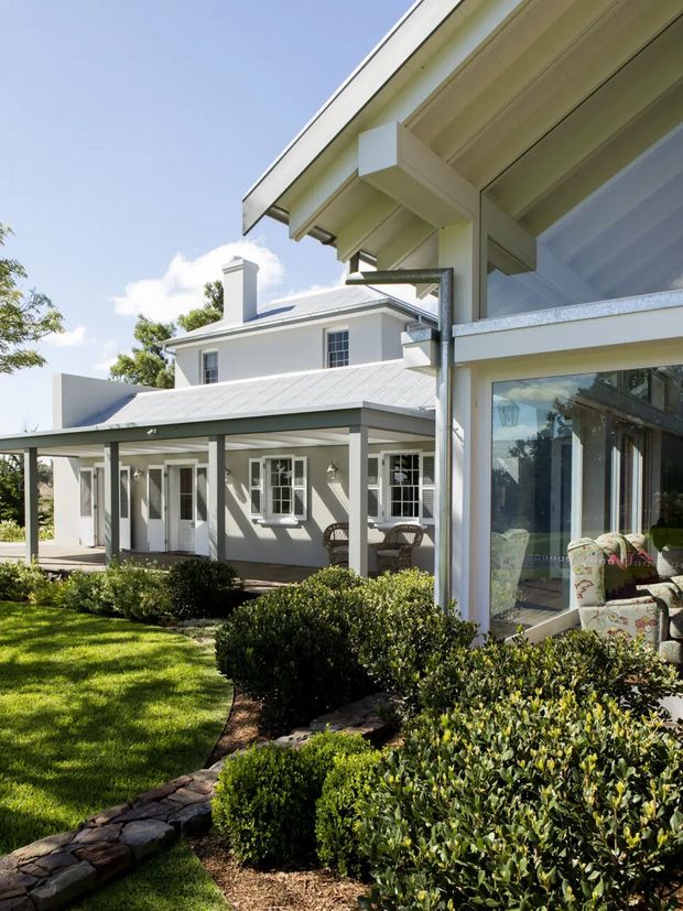 Sunlit exterior view of a refined light-coloured house with a corrugated metal roof, covered verandah, white-framed windows and doors, and a glazed extension or sunroom at right. Carefully clipped hedges, lawn, and stone edging form the foreground garden. The building has a traditional residential character with clearly expressed eaves and verandah structure.