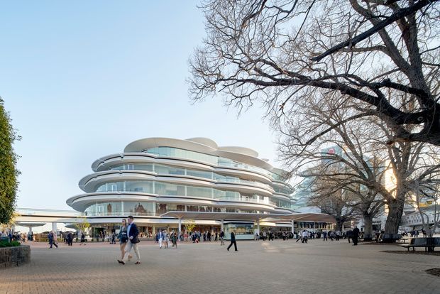 A wide exterior view of a distinctive multi-storey commercial building with rounded, layered floor plates, continuous curved glazing, and extensive balconies or terraces. The building fronts a broad paved public plaza with pedestrians, mature deciduous trees, and surrounding urban structures. The scene is bright and open, with a contemporary urban architectural character.