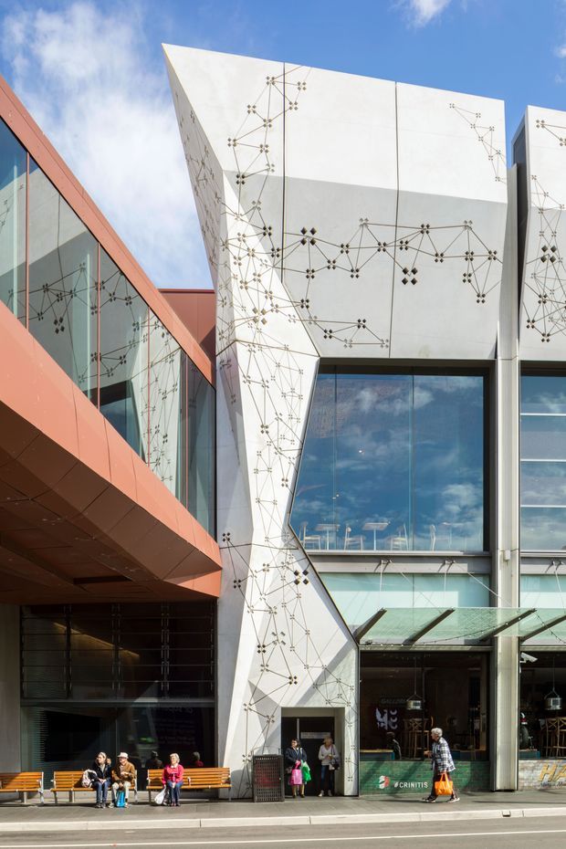 Exterior architectural photograph of a contemporary multi-storey commercial or civic building. The facade combines angular white panels decorated with an abstract network of dark geometric lines and nodes, large reflective glazed areas, and a bold terracotta-red projecting wing. Street-level shopfronts, entrances, benches, and pedestrians are visible, with a bright blue sky above.