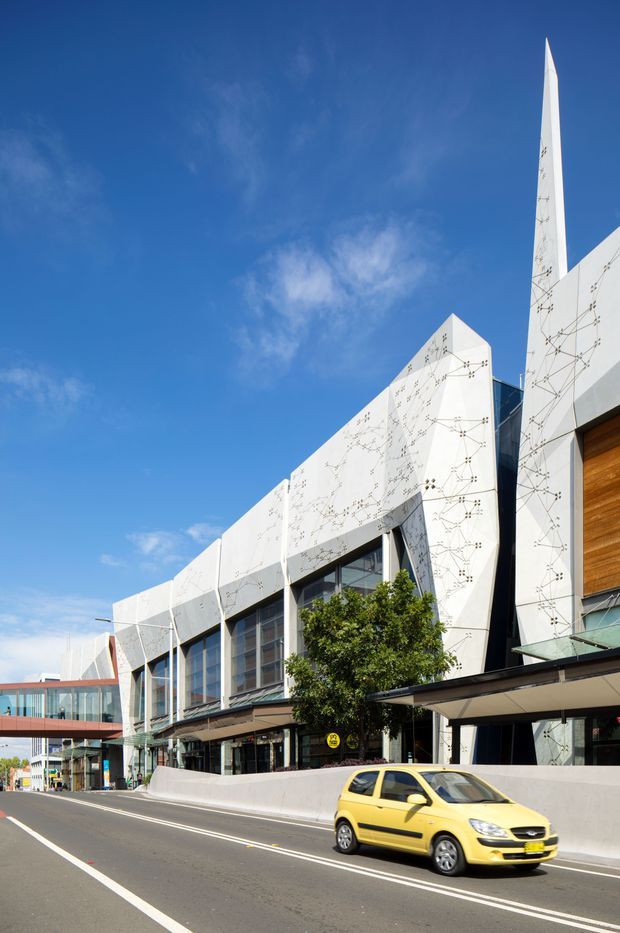 Street-level exterior view of a large contemporary urban building with a dramatic angular white facade, tall pointed vertical elements, expansive dark-framed glazing, and timber-clad detailing. The facade features an abstract fine-line geometric pattern. A covered entrance canopy and landscaped street edge are visible, with a city road in the foreground.