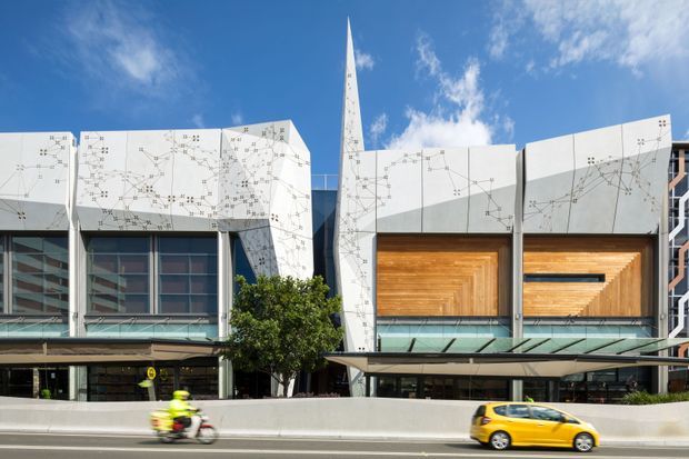 Street-facing view of a contemporary multi-storey commercial or institutional building with angular white facade panels covered in fine linework, large glazed sections, recessed timber-lined openings, and a tall pointed vertical architectural element. A covered entrance canopy and road barrier occupy the foreground.