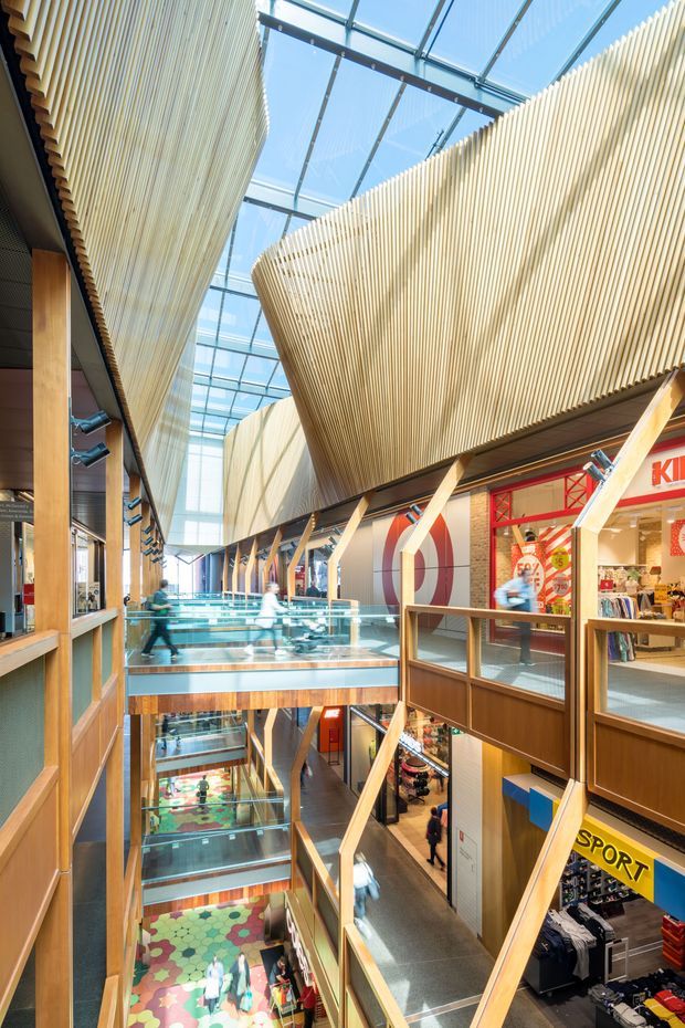 A bright multi-level commercial shopping interior with open walkways, glass balustrades, retail storefronts, timber structural elements, and dramatic suspended curved slatted timber screens beneath a large glazed roof. The atrium has strong natural daylight, visible escalator circulation, and a contemporary retail centre aesthetic.