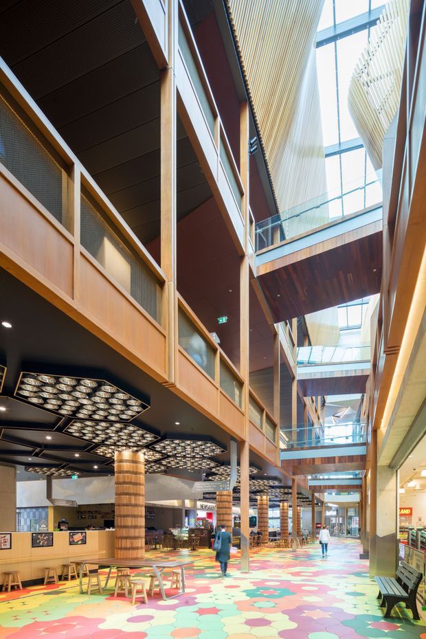 A tall, brightly lit multi-level commercial interior atrium with a central circulation space, extensive timber wall and ceiling finishes, exposed balconies and bridges, glass balustrades, clerestory glazing, and patterned geometric ceiling lights. The ground floor has colourful patterned flooring, seating, retail and dining areas, creating a lively public arcade or shopping-centre environment.