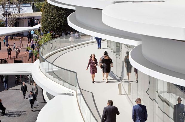 Exterior architectural view of a large multi-level commercial building with sweeping curved white floor plates, continuous glass balustrades, and extensive reflective glazing. Pedestrians move along elevated terraces and walkways, emphasizing the building’s layered circulation and sculptural contemporary form.