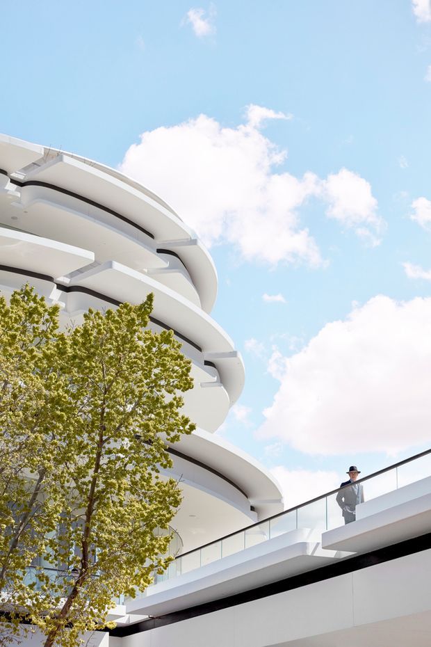 Upward-looking exterior architectural photograph of a multi-storey modern building with distinctive sweeping, curved white balconies and layered horizontal floor plates. A glass balustrade, black trim lines, and an integrated leafy tree are visible, with a person standing on an upper terrace. Bright blue sky and clouds form the background.