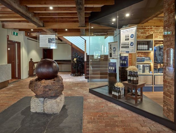 Interior view of a heritage-style commercial visitor centre or cellar entrance, with exposed timber beams, exposed brick walls, brick flooring, glass display cabinetry, informational signage, and branded bottles and merchandise. A sculptural stone-and-wood feature sits in the foreground, while stairs and exhibition elements lead further into the space.
