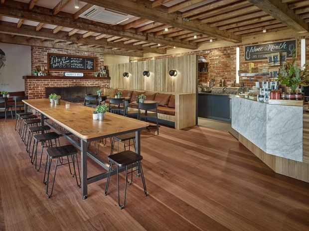 A warmly styled café or hospitality interior with a long communal timber dining table and black stools, banquette seating, exposed timber ceiling beams, exposed red-brick walls, timber flooring, and a compact service counter with a light veined stone-clad front. Chalkboards, shelving, pendant-free recessed lighting, plants, and coffee service details contribute to an industrial yet inviting atmosphere.