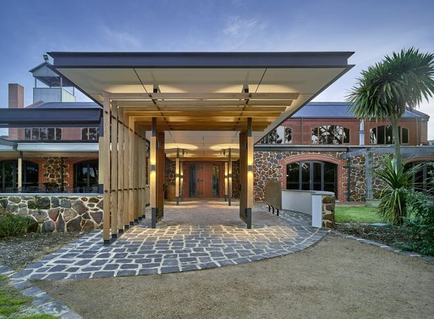 Exterior view of a substantial low-rise building with a broad flat-roofed entrance canopy supported by slender timber posts. The facade combines dark irregular stone masonry, extensive glazing, timber detailing, and reddish-brown brickwork. A curved stone-paved approach leads to the recessed timber entry door, with established palms and landscaped planting at the sides.