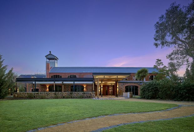 Twilight exterior view of a substantial low-rise building set behind a broad lawn and curving gravel or paved paths. The facade combines dark metal-framed glazing, exposed red brick, a long standing-seam metal roof, stone retaining walls, and a prominent glazed central entrance. A small tower-like rooftop structure and mature landscaping contribute to the character of the property.