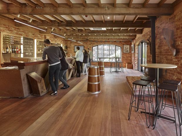A warm industrial-style bar or hospitality interior with exposed red brick walls, timber ceiling beams and joists, timber flooring, and a long service counter. High bar tables and stools occupy the right side, while patrons gather at the counter. Large arched and industrial-framed windows bring in daylight, and a barrel serves as a central feature.