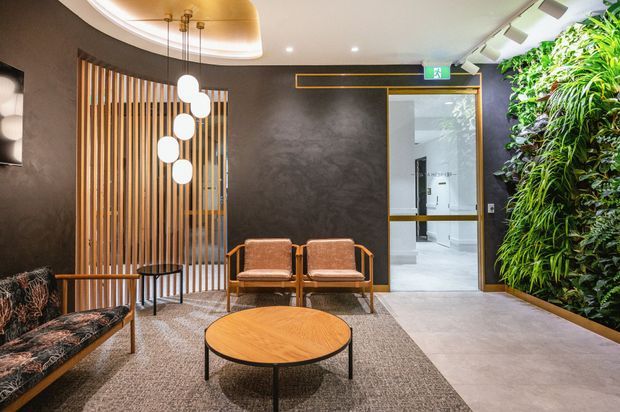 A refined contemporary reception or waiting area with dark textured walls, a curved recessed ceiling detail, timber slat screening, sculptural pendant lights, and mid-century-inspired upholstered lounge seating around a round timber coffee table. A lush vertical living wall lines the right side, while a glazed doorway opens to a bright tiled corridor or bathroom area.