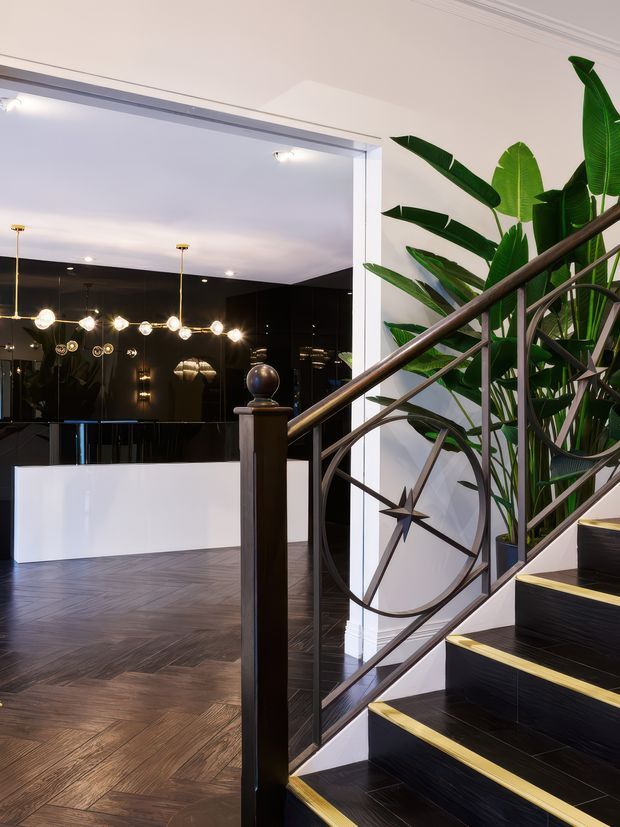 A refined interior stairway with dramatic black stairs edged in brass-gold trim, dark metal balustrading with a circular starburst motif, and a substantial dark timber newel post. Large tropical plants line the stair, while an open dining or living area beyond features dark glossy walls, a white half-height partition, and sculptural globe pendant lighting. The floor is laid in a dark herringbone timber pattern, creating a luxurious, high-contrast atmosphere.