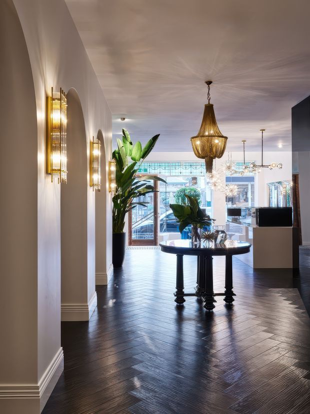 Elegant contemporary entry hall or foyer with a dark herringbone timber floor, white plaster walls, arched openings, ornate brass wall sconces, a large decorative pendant, tropical plants, and a round dark console table. Full-height glazing and a bright commercial-looking space are visible at the far end.