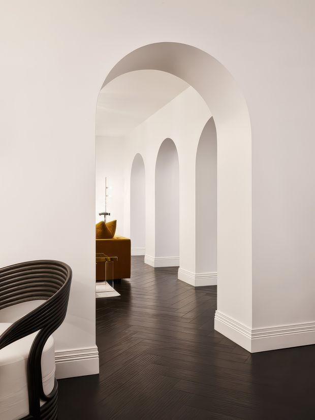 A refined interior hallway or passage framed by a large rounded arch, with a series of smaller arched openings beyond. The space has white plaster walls, ornate layered white baseboards, dark herringbone timber flooring, and a glimpse of a mustard-gold upholstered seating area. A sculptural black curved chair is partially visible in the foreground. The overall composition is minimalist, monochromatic, and strongly architectural.