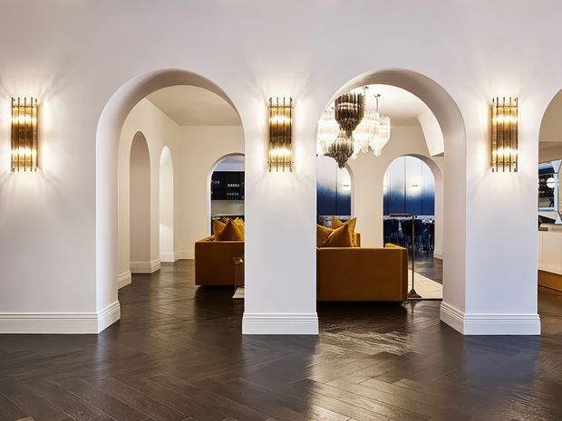 Elegant interior hallway or foyer with a series of rounded arched openings, white plaster walls, decorative wall sconces, dark herringbone timber flooring, and views into a warmly furnished living area and dining space. A mustard-gold sofa and ornate pendant chandeliers add a refined, luxurious character.
