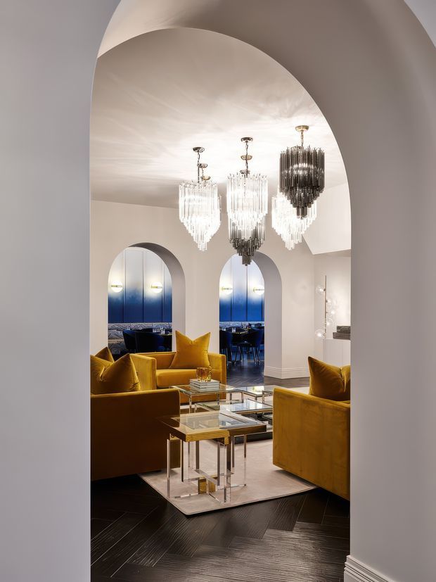A dramatic, refined living room viewed through a large rounded archway. The room features mustard-gold velvet sofas and armchairs, glass nesting coffee tables with metallic frames, a pale area rug, dark herringbone timber flooring, and several ornate cascading crystal chandeliers. Arched openings and mirrored or glossy blue-toned panels lead toward a dining or lounge area in the background.