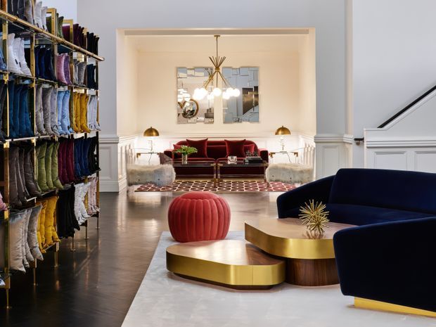 A richly styled interior showing a dramatic living room or lounge viewed from an open entry area. The space features dark timber flooring, white decorative wall panelling and column trim, a navy velvet sofa in the foreground, a deep red sofa at the rear, sculptural round coffee tables with brushed brass tops and dark timber bases, a red ribbed ottoman, a patterned rug, and a striking multi-arm chandelier. Shelving filled with folded textiles lines the left side, suggesting a design showroom or textile display area. The overall look is glamorous, refined, and Art Deco-inspired.