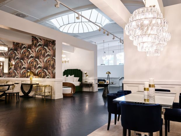 A refined open-plan interior combining dining and living areas. The space features dark herringbone timber flooring, white paneled walls, a dramatic botanical feature wallpaper, sculptural dark velvet dining chairs, a marble-topped dining table, brass accents, and a large tiered crystal chandelier. A curved skylight and exposed track lighting add architectural interest, while a green upholstered feature wall and contemporary furnishings create a polished, luxurious atmosphere.