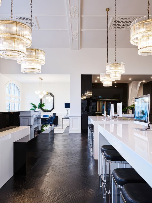 A refined, high-end hospitality interior combining a bright bar and reception area with a formal lounge or dining zone. The space features glossy white counters, black cabinetry, black leather bar stools, dark herringbone timber flooring, ornate white wall panelling and ceiling detailing, arched windows, and multiple tiered crystal chandeliers suspended from the ceiling. The palette is predominantly white, black, and dark timber with polished metallic accents.