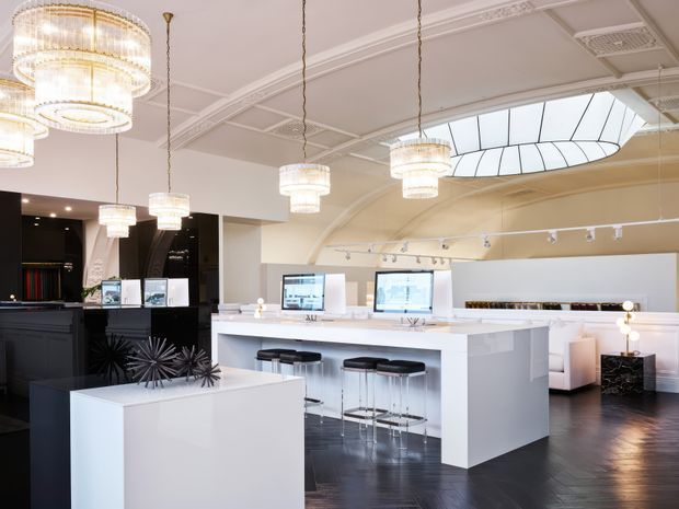 Bright, upscale commercial interior with a white reception or service counter, integrated workstations, bar stools, black display cabinetry, dark timber flooring, crystal pendant chandeliers, decorative ceiling mouldings, and a large curved skylight. The space has a polished showroom, hospitality, or salon-like character.
