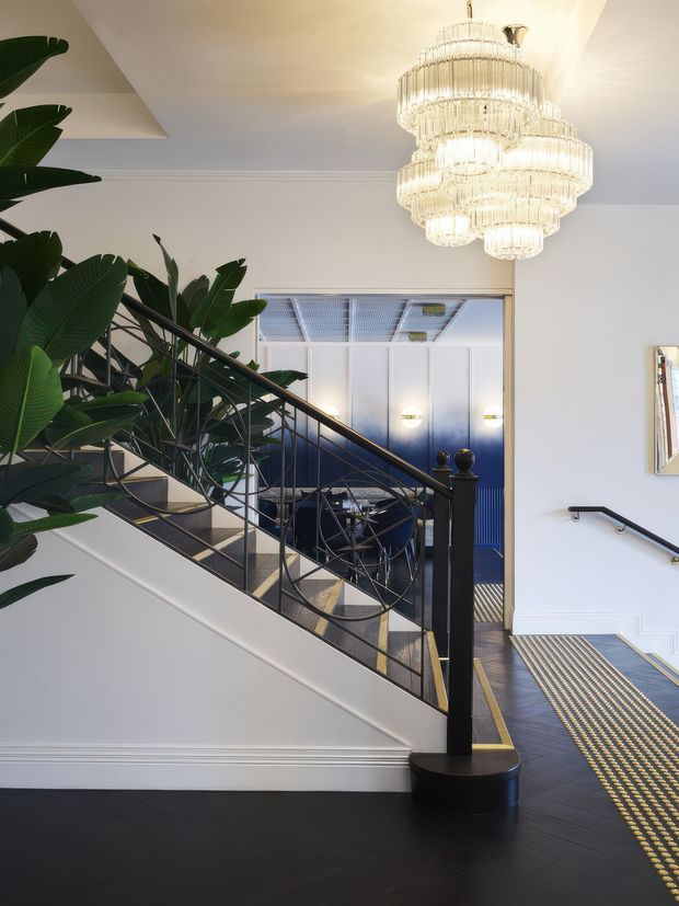 An elegant interior stairway and landing with dark timber flooring, white paneled walls, black metal balustrades, and a large tiered crystal chandelier. A dramatic leafy indoor plant fills the stairwell, while a dining or lounge area is visible beyond a wide opening with blue-toned cabinetry or wall panels. Decorative tactile flooring strips lead along the landing, and a wall-mounted handrail continues toward the adjoining space.
