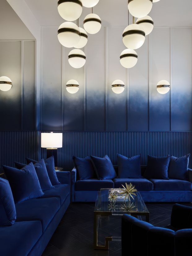 A dramatic, luxurious living room in a deep navy and blue palette. Multiple sculptural globe pendant lights hang in front of a white-to-navy gradient wall with vertical panel detailing and a dark ribbed lower wall. Several plush navy sofas and armchairs surround a glass coffee table with a metallic starburst sculpture, while a table lamp adds a warm glow. The space has a moody, high-contrast atmosphere with reflective and geometric detailing.