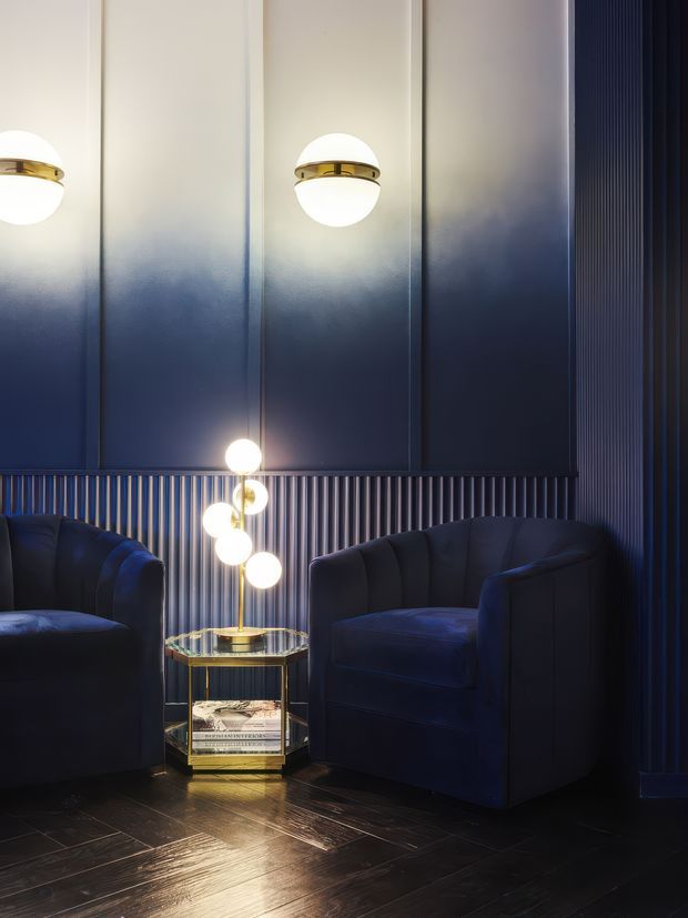 A moody, luxurious lounge corner with two deep navy upholstered armchairs, a small round glass-and-gold side table, and a sculptural globe-bulb lamp. The dark blue paneled and vertically fluted wall treatment, reflective dark timber floor, and warm metallic details create a dramatic art deco-inspired interior.