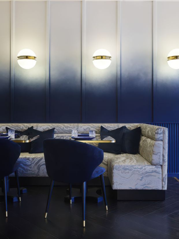 A sophisticated restaurant or hospitality dining area with a curved upholstered banquette, navy velvet dining chairs, small round tables set with glassware and tableware, and dramatic spherical wall sconces with brass bands. The room features dark blue gradient wall panelling, patterned upholstery, and dark tiled flooring, creating a moody, luxurious atmosphere.
