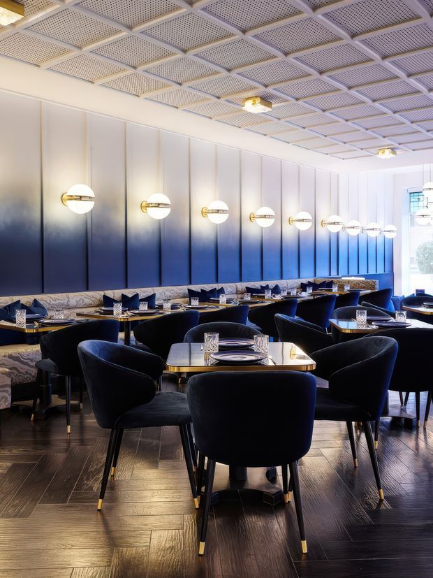 A sophisticated restaurant dining room with rows of elegantly set tables and deep navy upholstered dining chairs. The space features a dramatic gradient wall treatment transitioning from pale grey to rich blue, evenly spaced globe wall sconces with brass-toned fittings, dark timber flooring, patterned banquette seating, and a distinctive coffered ceiling with perforated panels. The overall atmosphere is refined, moody, and luxurious.