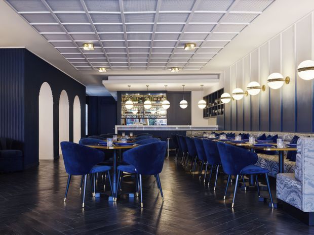 A sophisticated restaurant dining room with a dark navy and blue colour scheme. The space features rows of blue velvet dining chairs, patterned upholstered banquette seating, dark timber flooring, a long bar or service counter at the rear, and a striking linear arrangement of spherical pendant lights. A white modular grid ceiling contrasts with dark fluted wall panels and arched wall recesses, creating a polished contemporary hospitality interior.