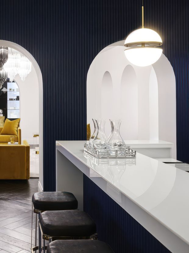 A sophisticated interior view of a glossy white kitchen island or bar counter with black upholstered stools, set against deep navy fluted wall panelling. A sculptural white pendant light hangs above, while large arched openings reveal a refined living area with mustard seating and decorative lighting. The space combines strong geometric arches, reflective surfaces, and high-contrast navy, white, black, and gold detailing.