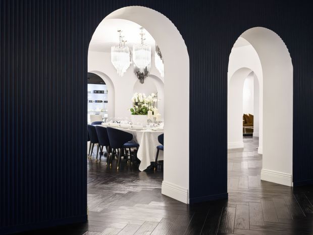 Elegant interior with deep navy fluted wall panelling framing two large rounded archways. The central opening reveals a formal dining room with a white tablecloth, dark upholstered dining chairs, chandeliers, and floral arrangements, while the adjoining opening leads toward a bright white hallway or living area. Dark herringbone timber flooring, white arched plasterwork, and refined decorative detailing create a dramatic, luxurious atmosphere.