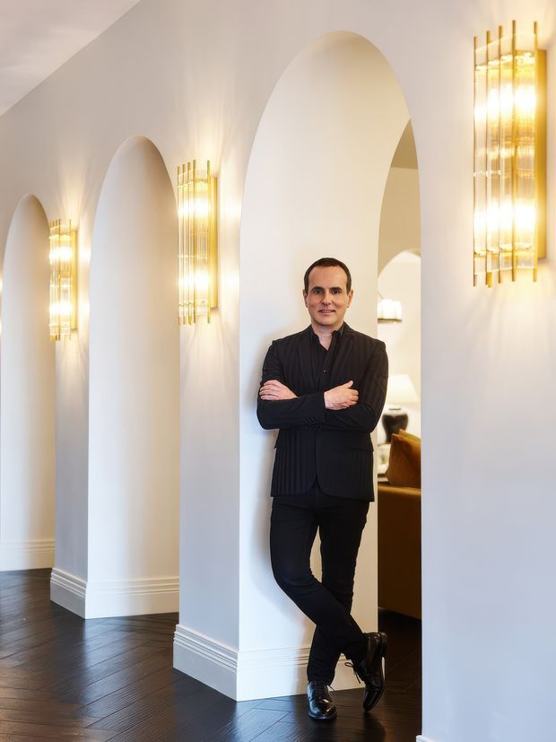 A refined interior hallway with a series of tall rounded arch openings and white plaster walls, illuminated by elegant vertical gold-toned wall sconces. Dark timber flooring and decorative white skirting create a high-contrast, polished setting. A person stands in the central archway, with a glimpse of a furnished room beyond.