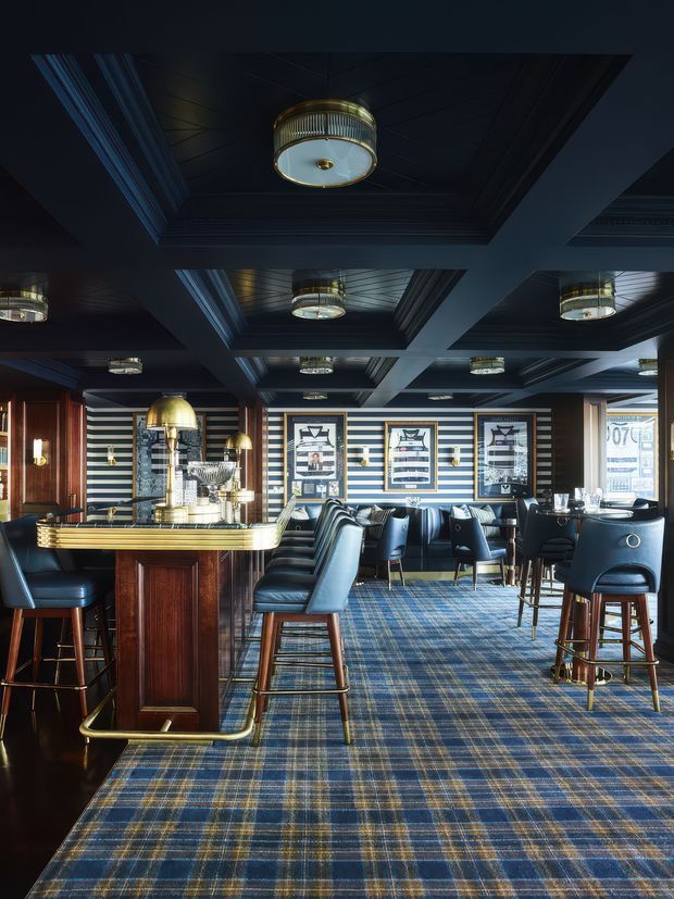 A sophisticated bar and dining lounge interior with dark navy coffered ceilings, multiple brass and glass pendant lights, striped navy-and-white wall finishes, framed artwork, dark timber bar cabinetry, leather bar stools and dining chairs, and a blue plaid carpet. The space has a classic, moody hospitality-club atmosphere with polished brass detailing and warm timber accents.