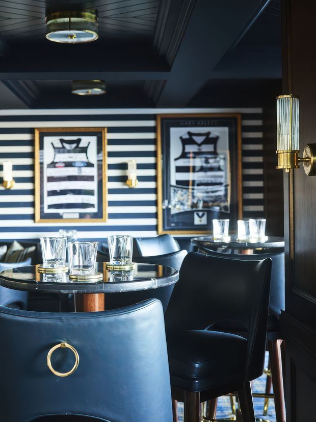 A compact, sophisticated home bar or lounge with dark navy and black finishes. The room features striped navy-and-white wall treatment, framed artwork, glossy dark ceiling panels, brass wall sconces and pendant lighting, and black leather bar chairs arranged around small round tables. Crystal tumblers and decanters reinforce the refined cocktail-bar atmosphere.