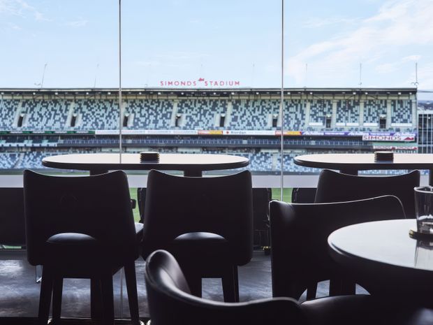 A refined hospitality or dining interior with dark upholstered chairs and round tables arranged beside floor-to-ceiling glazing. The windows overlook a large sports stadium with tiered seating and a visible playing field, creating a dramatic indoor-outdoor connection. The space has a dark, contemporary fit-out with strong natural daylight and panoramic views.