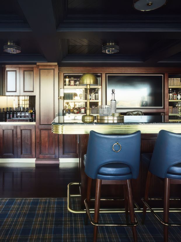 A richly detailed, dark-toned home bar with deep timber wall panelling, a brass-trimmed bar counter, built-in bottle storage, illuminated glass shelving, and two deep blue upholstered bar stools. The coffered ceiling has dark painted beams and integrated decorative lighting, while a large reflective screen or mirror sits behind the bar. A tartan-patterned carpet adds a tailored accent.