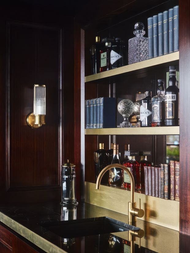 A dark, richly finished home bar with built-in open shelving displaying blue-bound books, liquor bottles and crystal decanters. The cabinetry and wall panelling are deep dark timber, with brushed brass shelf trims, a brass wall sconce, and a brass gooseneck tap over a black sink and dark benchtop. The overall look is elegant, moody and highly tailored.