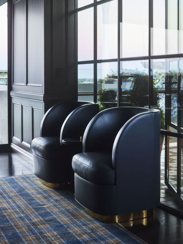 A refined interior seating area with two sculptural black leather barrel armchairs positioned beside large black-framed glazed windows. The room features dark decorative wall panelling, polished dark flooring, brass-toned chair bases, and a blue, grey, and beige plaid rug. Natural light enters through the expansive glazing, with greenery and an outdoor view visible beyond.