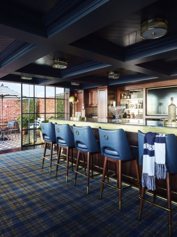 A richly styled home bar with a long dark timber and stone counter, upholstered blue bar stools, extensive built-in shelving, glassware and decorative objects. The ceiling features dramatic dark coffered panels with blue-painted trim and multiple cylindrical pendant lights. A glazed folding or sliding wall opens to a brick-paved outdoor terrace, while a blue patterned carpet and warm timber joinery create a luxurious, club-like atmosphere.