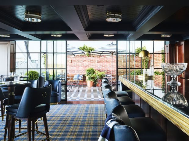 A refined bar and lounge interior with a long polished counter, black leather bar stools, dark upholstered dining chairs, and a blue-and-gold patterned carpet. Large black-framed glazed doors and windows open onto a brick-walled terrace with outdoor seating. The dark coffered ceiling contains multiple decorative drum and recessed light fixtures, while brass and glass decorative elements add a luxurious, classic feel.