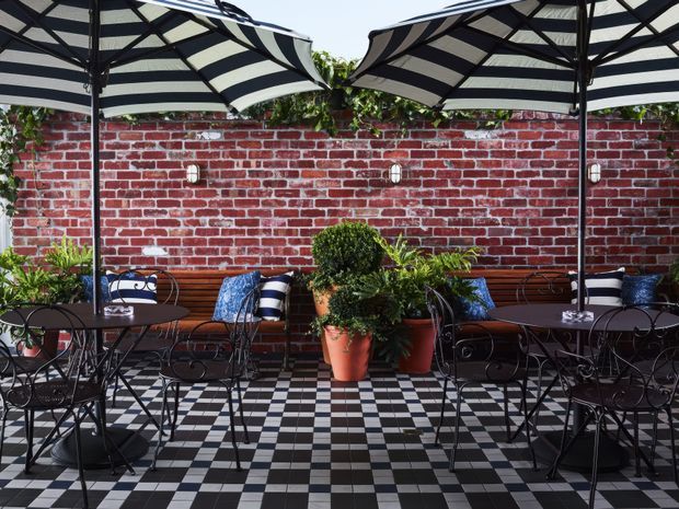 A stylish outdoor courtyard dining terrace with black-and-white checkerboard flooring, a red brick boundary wall, wrought-iron café tables and chairs, striped umbrellas, bench seating, and potted topiary and plants. The space has a refined, European-inspired café atmosphere.