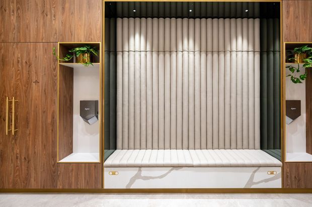 A symmetrical built-in upholstered bench or booth set into a richly grained timber wall. The seating features vertical channel tufting in a pale neutral fabric, a matching cushioned seat, dark recessed overhead backing with spotlights, white marble-look base panels, and integrated display niches with small plants and fixtures on either side.