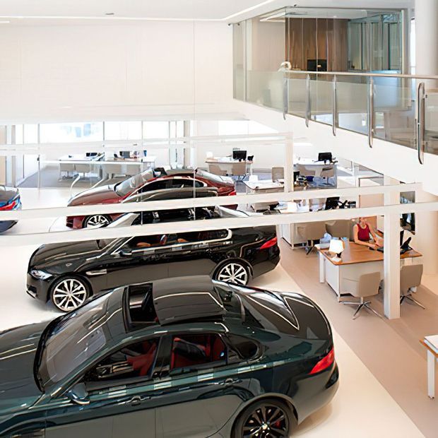 Bright, multi-level automotive showroom with several luxury cars displayed on a polished light floor. White structural framing, extensive glazing, open-plan sales desks, and an upper-level glass-balustraded mezzanine create a spacious contemporary commercial environment.