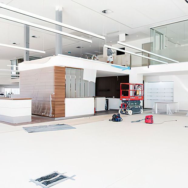 Bright, open-plan contemporary interior under construction or renovation, featuring white walls and ceilings, extensive glazing and mirrored surfaces, exposed installation equipment, a compact kitchen or service joinery area with white cabinetry and warm timber panelling, and an upper-level mezzanine or balustrade.